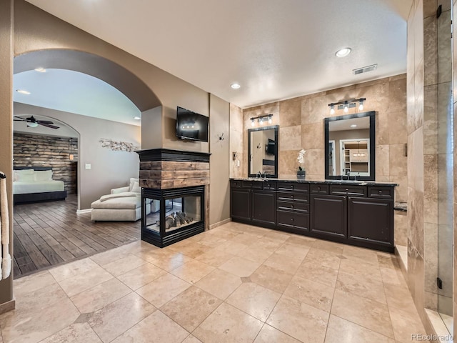 full bath featuring ceiling fan, a multi sided fireplace, visible vents, tile walls, and double vanity