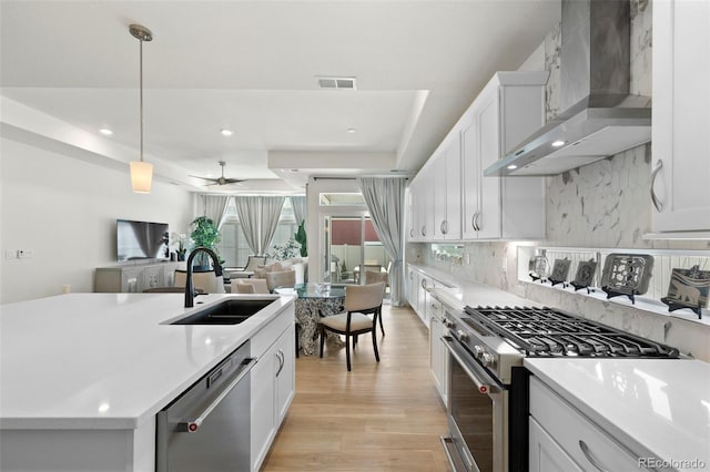 kitchen featuring wall chimney range hood, decorative backsplash, light wood-style flooring, appliances with stainless steel finishes, and a sink