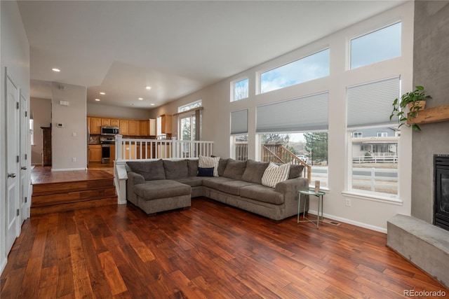 living room with dark wood-style flooring, a glass covered fireplace, a towering ceiling, and baseboards