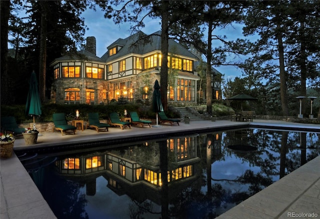 back of property at dusk featuring a patio area, stone siding, an outdoor pool, and a chimney