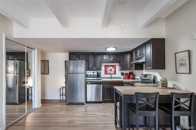 kitchen with light wood finished floors, under cabinet range hood, beamed ceiling, stainless steel appliances, and a sink