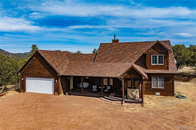 chalet / cabin with driveway, an attached garage, a shingled roof, a chimney, and a patio area