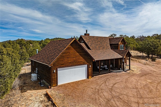 chalet / cabin with a shingled roof, dirt driveway, covered porch, a chimney, and a garage