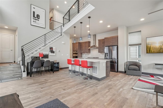 kitchen with decorative light fixtures, an island with sink, a kitchen breakfast bar, stainless steel refrigerator with ice dispenser, and wall chimney range hood