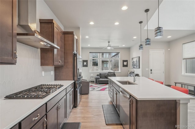 kitchen featuring pendant lighting, sink, a kitchen island with sink, stainless steel appliances, and wall chimney exhaust hood