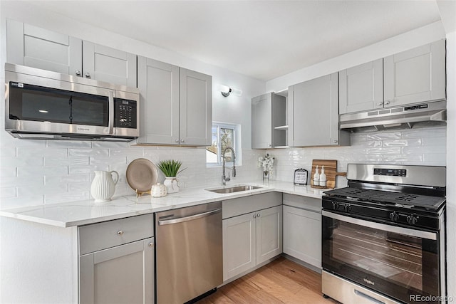 kitchen featuring stainless steel appliances, under cabinet range hood, a sink, gray cabinetry, and tasteful backsplash