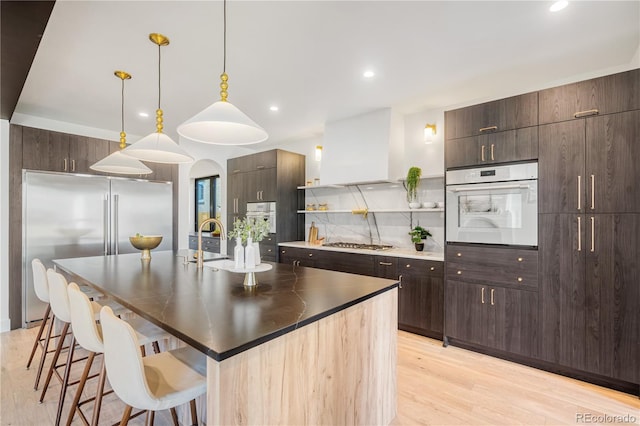 kitchen with appliances with stainless steel finishes, exhaust hood, a center island with sink, light hardwood / wood-style flooring, and hanging light fixtures