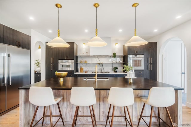 kitchen featuring decorative light fixtures, dark brown cabinetry, stainless steel appliances, and a kitchen island with sink