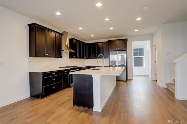 kitchen with sink, an island with sink, stainless steel appliances, light hardwood / wood-style floors, and wall chimney range hood