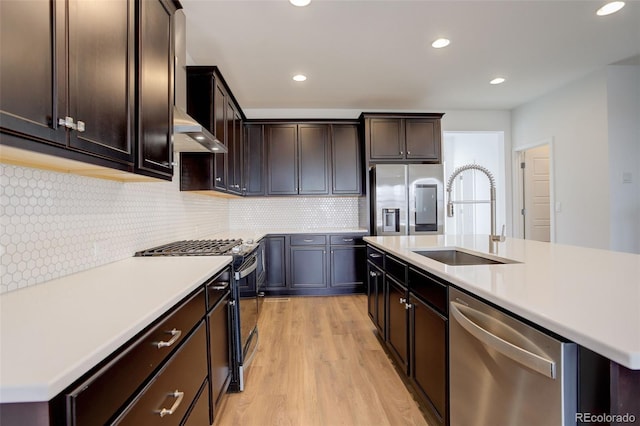 kitchen featuring appliances with stainless steel finishes, sink, wall chimney exhaust hood, a center island with sink, and light hardwood / wood-style flooring