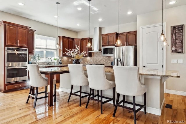 kitchen featuring light wood-style floors, appliances with stainless steel finishes, wall chimney range hood, a large island, and decorative backsplash