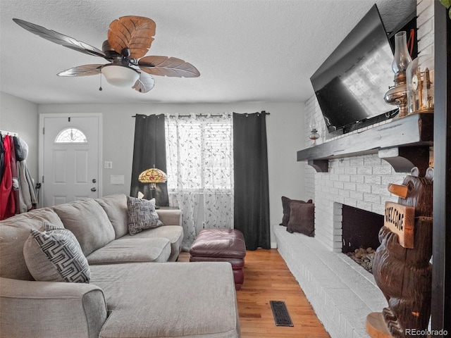 living room with a textured ceiling, light wood-style flooring, visible vents, a ceiling fan, and a brick fireplace