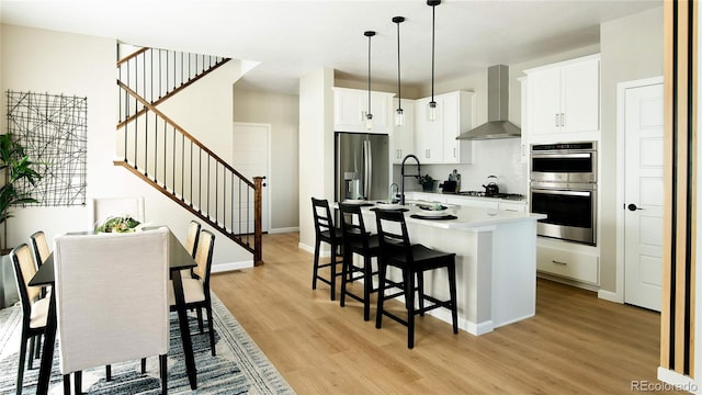 kitchen featuring wall chimney range hood, appliances with stainless steel finishes, decorative backsplash, light wood finished floors, and a kitchen bar