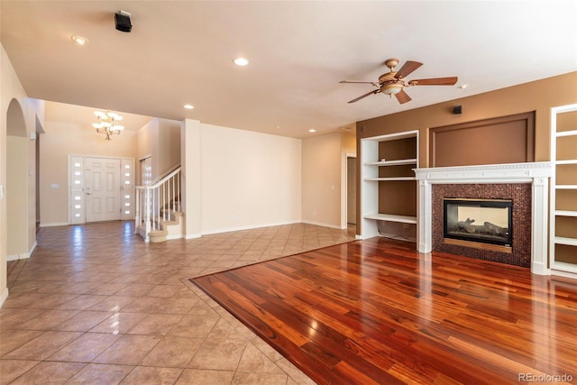 unfurnished living room with a multi sided fireplace, ceiling fan with notable chandelier, built in features, and light tile patterned flooring