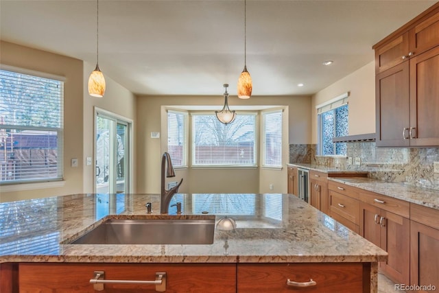 kitchen featuring a kitchen island with sink, sink, hanging light fixtures, decorative backsplash, and light stone counters