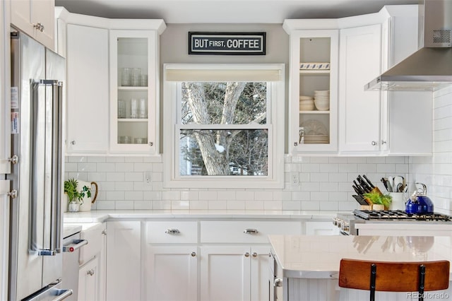 kitchen featuring white cabinets, light countertops, appliances with stainless steel finishes, wall chimney range hood, and glass insert cabinets