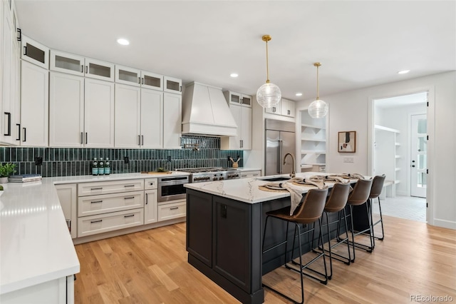 kitchen featuring premium range hood, a center island with sink, white cabinets, built in appliances, and light hardwood / wood-style floors