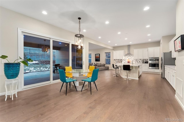 kitchen featuring decorative light fixtures, wall chimney range hood, an island with sink, stainless steel appliances, and white cabinets