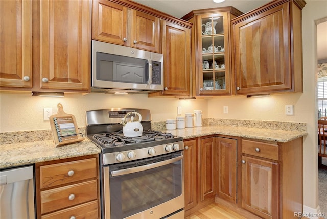 kitchen with brown cabinets, glass insert cabinets, appliances with stainless steel finishes, and light stone countertops