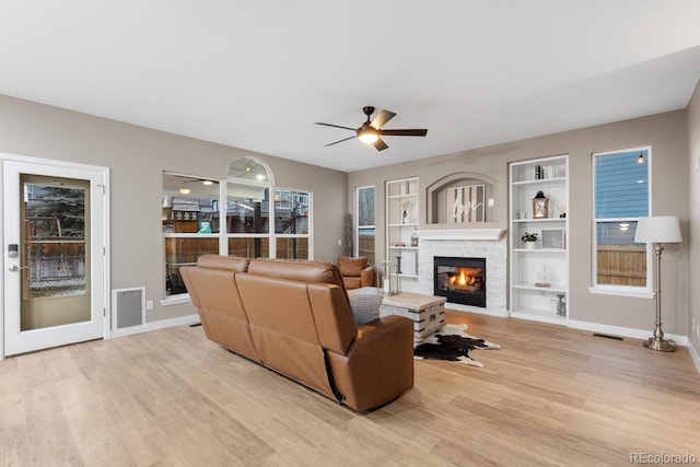 living room with baseboards, built in features, light wood-style flooring, a glass covered fireplace, and a ceiling fan