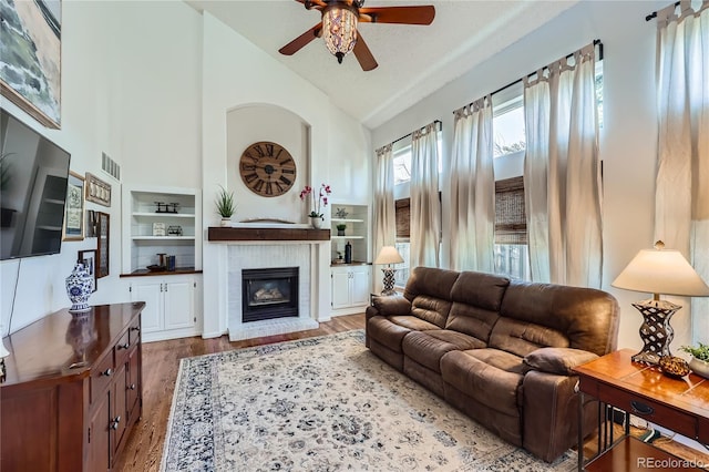 living room with high vaulted ceiling, a fireplace, ceiling fan, light hardwood / wood-style floors, and built in shelves