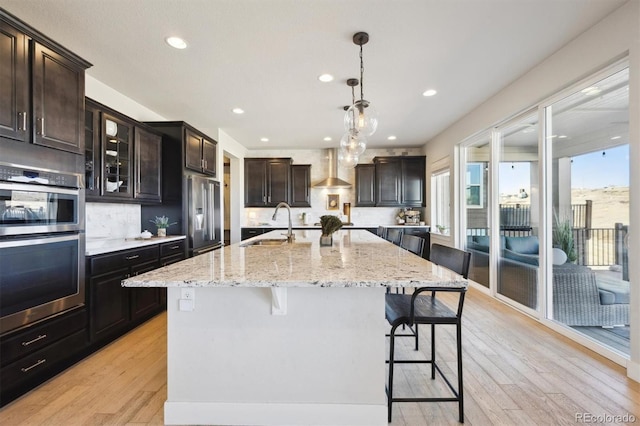 kitchen with appliances with stainless steel finishes, light wood finished floors, wall chimney exhaust hood, and a kitchen bar