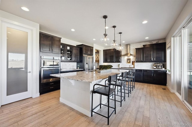 kitchen with a kitchen breakfast bar, wall chimney range hood, appliances with stainless steel finishes, light wood finished floors, and tasteful backsplash