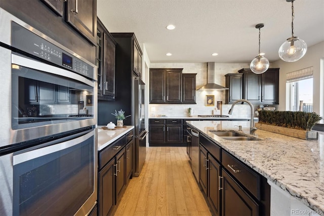 kitchen featuring hanging light fixtures, appliances with stainless steel finishes, a sink, wall chimney range hood, and light wood-type flooring