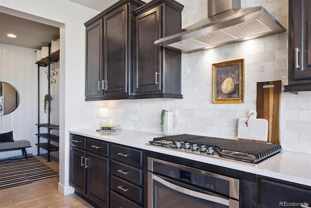 kitchen featuring stainless steel appliances, light wood-style floors, light countertops, wall chimney exhaust hood, and tasteful backsplash