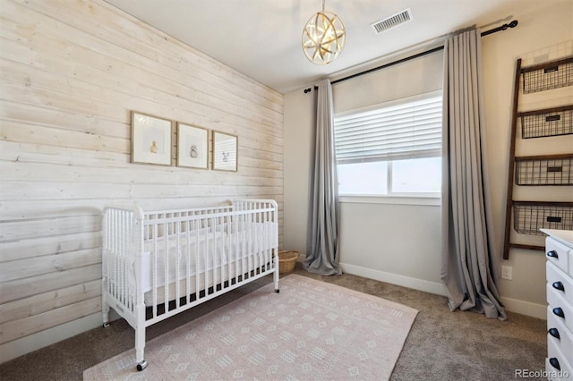 carpeted bedroom with visible vents, an inviting chandelier, wooden walls, a nursery area, and baseboards