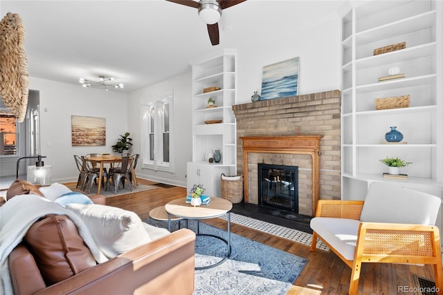 living room with dark wood-type flooring, ceiling fan, built in shelves, and a brick fireplace