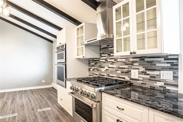 kitchen featuring wall chimney range hood, tasteful backsplash, lofted ceiling with beams, dark stone counters, and appliances with stainless steel finishes