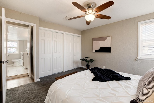 bedroom featuring two closets, ensuite bath, dark colored carpet, ceiling fan, and a textured wall