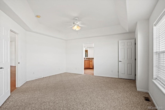 unfurnished bedroom with a raised ceiling, ceiling fan, light carpet, and ensuite bath