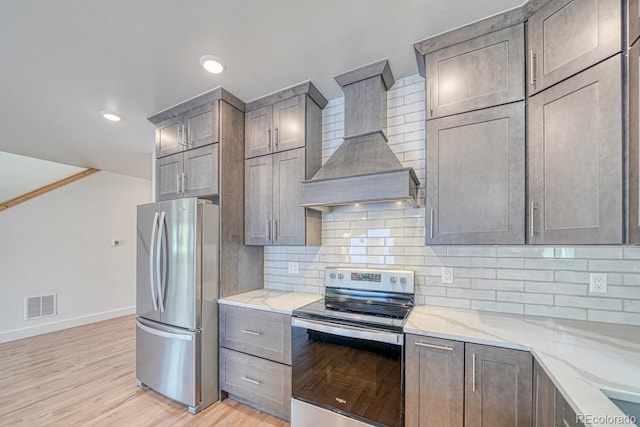 kitchen featuring backsplash, premium range hood, appliances with stainless steel finishes, light hardwood / wood-style floors, and light stone counters