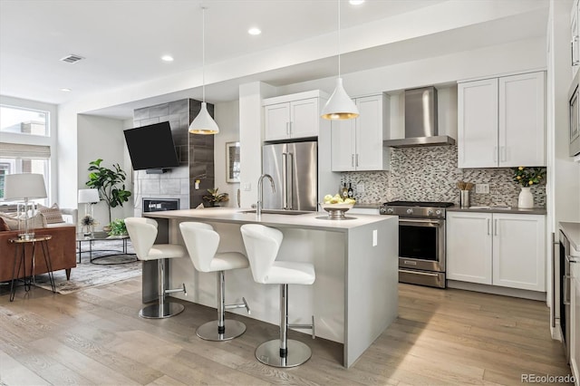 kitchen with wall chimney exhaust hood, a center island with sink, high end appliances, and decorative light fixtures