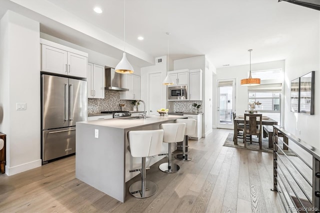 kitchen with white cabinets, appliances with stainless steel finishes, wall chimney range hood, a kitchen island with sink, and a breakfast bar area