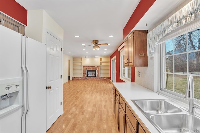 kitchen with white refrigerator with ice dispenser, brown cabinetry, a brick fireplace, a sink, and light wood-type flooring