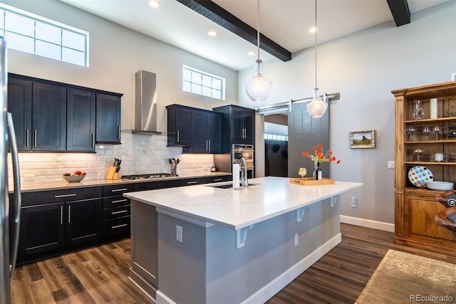 kitchen featuring wall chimney exhaust hood, hanging light fixtures, a center island with sink, beam ceiling, and appliances with stainless steel finishes