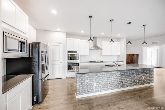 5651 Gore Creek Pl, Brighton, CO 80601 - kitchen featuring appliances with stainless steel finishes, wall chimney range hood, white cabinetry, light wood-style flooring, and a kitchen island with sink