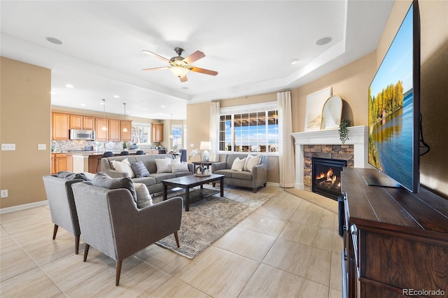 living room featuring ceiling fan, a raised ceiling, a stone fireplace, and light tile patterned flooring