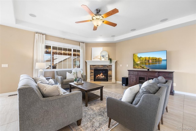 living room featuring ceiling fan, a stone fireplace, light tile patterned flooring, and a tray ceiling