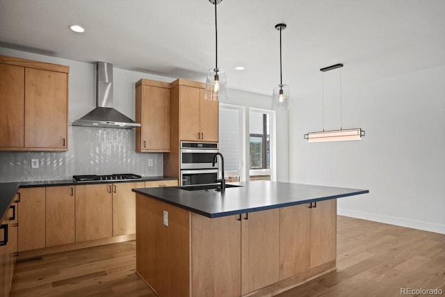 kitchen featuring decorative light fixtures, a center island with sink, appliances with stainless steel finishes, wall chimney range hood, and backsplash