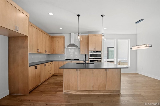 kitchen with light brown cabinetry, an island with sink, sink, hanging light fixtures, and wall chimney range hood