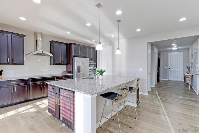 kitchen with wall chimney range hood, light hardwood / wood-style flooring, a kitchen island with sink, and appliances with stainless steel finishes