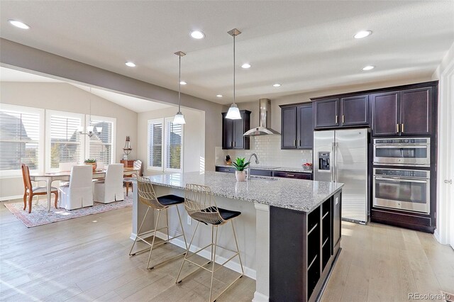 kitchen with wall chimney range hood, a center island with sink, vaulted ceiling, light wood-type flooring, and appliances with stainless steel finishes