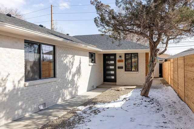 snow covered property entrance featuring crawl space, brick siding, fence, and roof with shingles