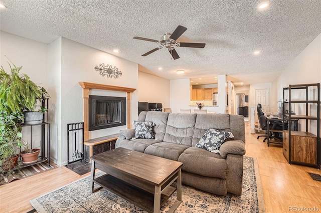 living area featuring a fireplace with raised hearth, a ceiling fan, light wood-style flooring, a textured ceiling, and recessed lighting