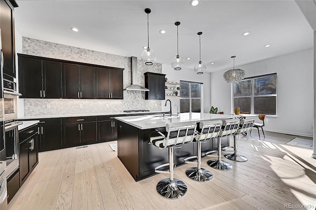 kitchen featuring a center island with sink, wall chimney exhaust hood, a breakfast bar, light countertops, and light wood-style floors