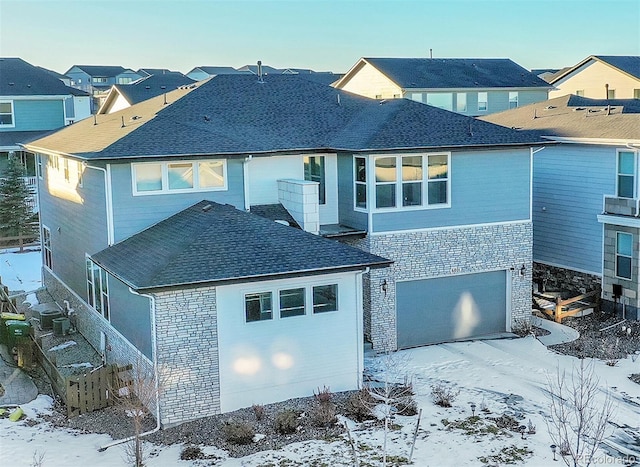 snow covered rear of property with stone siding, roof with shingles, an attached garage, and a residential view